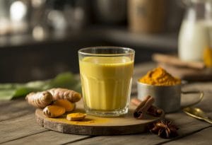 A glass cup of golden turmeric milk on a wooden table surrounded by turmeric roots, turmeric powder, and spices.
