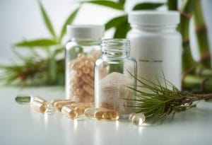 Close-up of silica supplement capsules and glass bottles surrounded by horsetail plant sprigs and bamboo leaves on a white surface.
