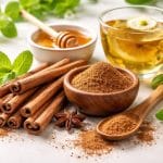 Close-up of cinnamon sticks and ground cinnamon arranged with green leaves, honey, and herbal tea on a white surface.